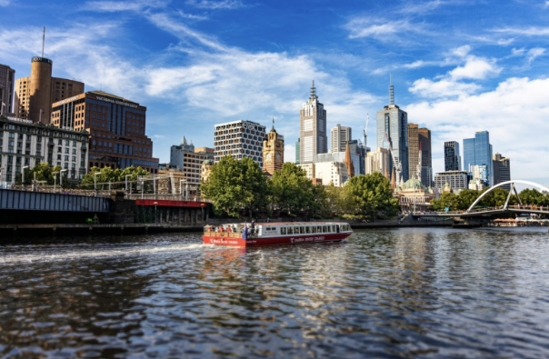 Melbourne city skyline featuring a red Yarra River Cruise boat on the river with skyscrapers and lush greenery in the background under a bright blue sky.